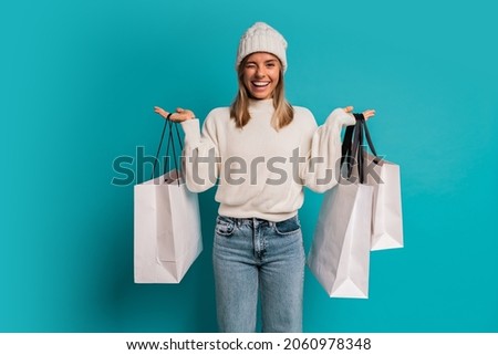 Studio winter portrait of  smiling blond woman in white hat and whool sweater  a holding  shopping bags ,posing in studio over turquose background. 