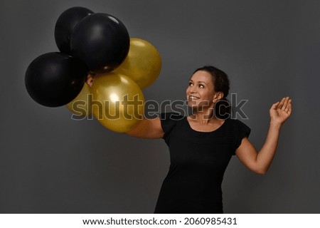Cheerful pretty woman with beautiful smile poses with inflated black and gold air balloons against gray wall background with copy space. Concept of Black Friday and gold credit card for shopping
