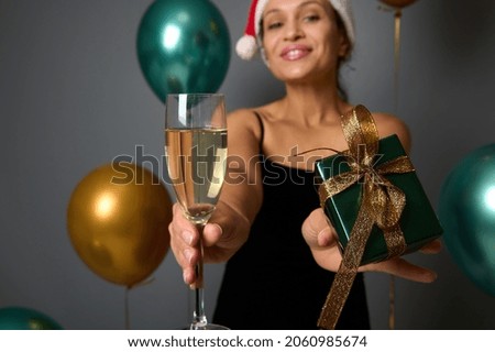 Focus in champagne glass and Christmas gift in shiny luxury wrapping paper with golden bow in the hands of cheerful woman in Santa hat celebrating New Year at party on gray background with copy space