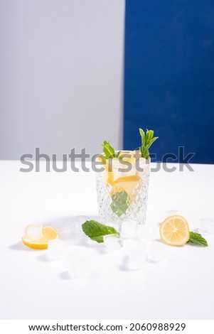 creative summer cocktail with ice in a glass glass with lemon, mint and ice cubes on a white-blue background.