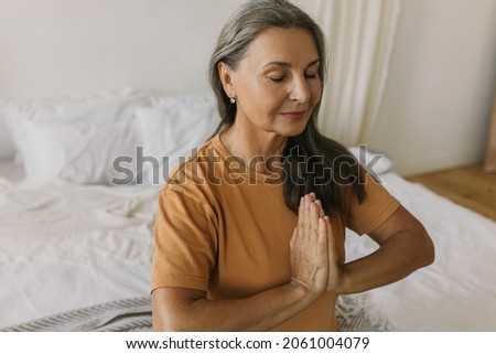 Upper view of pretty grey-haired mature woman in yellow t-shirt meditating at home sitting on white unmade bed reaching zen, pressing palms together in namaste gesture. Yoga, spiritual practice