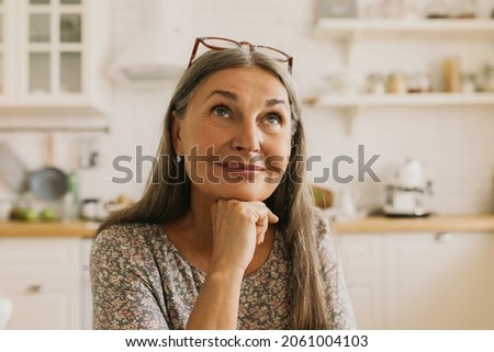 Charming lady of 60s surrounded with kitchen interior relaxing and dreaming, trying to decide what to cook for dinner, wearing glasses on forehead looking elegant in casual flowered dress Royalty-Free Stock Photo #2061004103