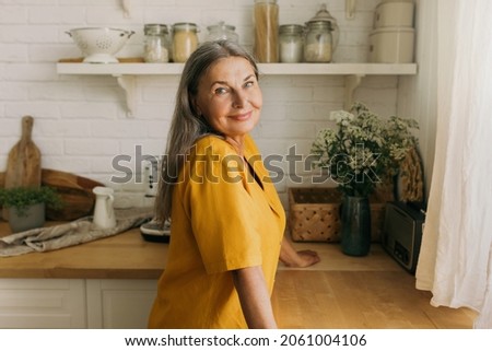 Lovely senior woman in yellow dress posing in countryhouse. Smiling positive female going to cook on kitchen, look from window. Weekends away, nature, interior, pure joy, growing old concept Royalty-Free Stock Photo #2061004106