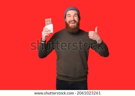 Happy bearded man is enjoying chocolate and showing like gesture at the camera. Photo over red background.