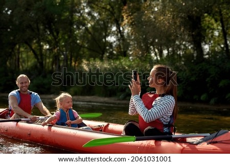Woman taking picture of husband and daughteron smartphone during floating on kayak. Concept of rest, leisure and weekend. Extreme water sport. Young caucasian family wearing life vests. Sunny daytime