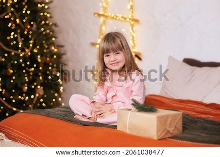 Happy little smiling girl in pajamas with gift on xmas Eve lies on bed. child opens New year gift at home near christmas tree with glowing lights garland. Kid sleep in bed in child bedroom at home. 