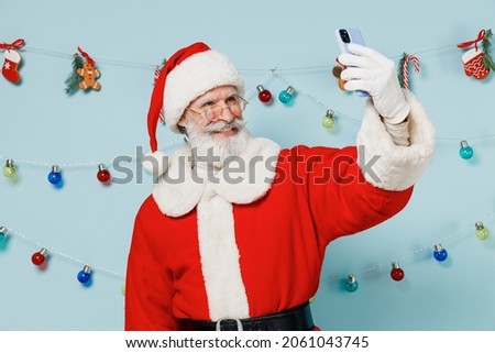 Old bearded Santa Claus man 50s wears Christmas hat red suit doing selfie shot on mobile cell phone isolated on plain blue background studio. Happy New Year 2022 celebration merry ho x-mas concept.