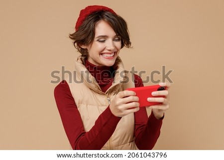 Young gambling woman wears red turtleneck vest beret using play racing app on mobile cell phone hold gadget smartphone for pc video games isolated on plain pastel beige background studio portrait.