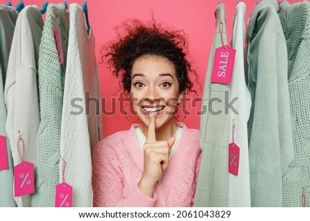 Young happy costumer woman 20s in sweater stand near clothes rack with tag sale in store showroom say hush be quiet with finger on lips shhh gesture isolated on plain pink background studio portrait