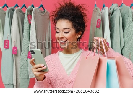 Young costumer fun woman in sweater stand near clothes rack with tag sale in store showroom hold package bags with purchases after shopping mobile cell phone isolated on plain pink background studio