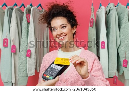 Young woman in sweater stand near clothes rack with tag sale in store showroom hold wireless modern bank payment terminal process acquire credit card payments isolated on plain pink background studio