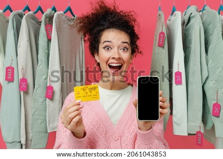 Young shocked woman in sweater stand near clothes rack with tag sale in store hold mobile cell phone blank screen workspace area credit bank card shop online isolated on plain pink background studio
