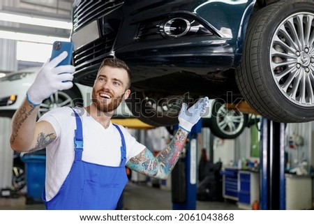Young fun professional technician mechanic man in blue overalls t-shirt do selfie shot on mobile cell phone stand near car lift check technical condition work in vehicle repair shop workshop indoors