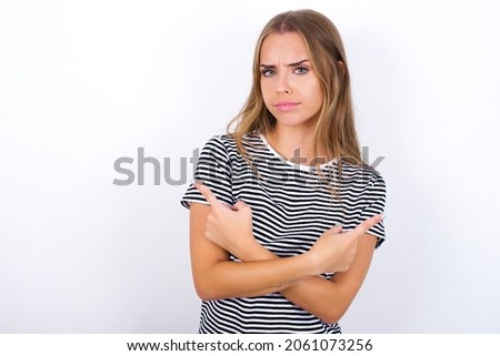 Serious beautiful blonde girl wearing striped t-shirt on white background crosses hands and points at different sides hesitates between two items. Hard decision concept