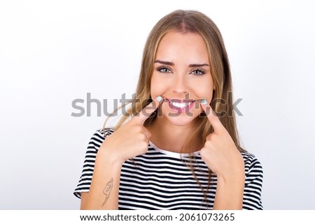 Happy beautiful blonde girl wearing striped t-shirt on white background with toothy smile, keeps index fingers near mouth, fingers pointing and forcing cheerful smile