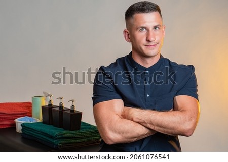 Portrait of smiling masseur with crossed arms. Career as a massage therapist or heropractor. Heropractor next to massage supplies. Massage room employee on gray background. Heropractic worker.