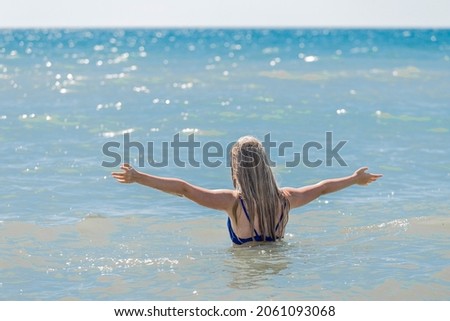 A young teenage girl with long blonde hair raised and spread her arms to the sides against the background of the sea water and horizon line. The concept of freedom