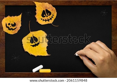 Yellow leaves with Halloween faces on the chalkboard, the girl's hand writes in white chalk. Copy space for text. Flat lay