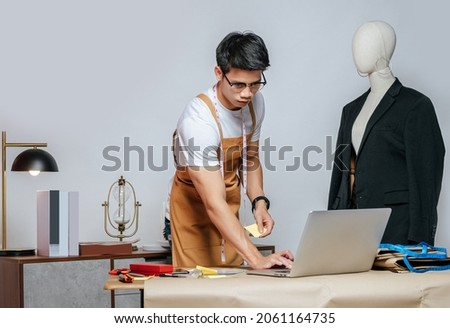 Portrait young Asian male fashion designer or tailor wearing eyeglasses use laptop computer to search idea or online orders, mannequin in suit and full of tailoring tools on desk in studio