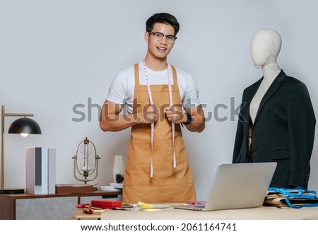 Cheerful young Asian fashion designer or tailor wearing eyeglasses use laptop computer to search idea or online orders, mannequin in suit and full of tailoring tools with equipment on desk in studio