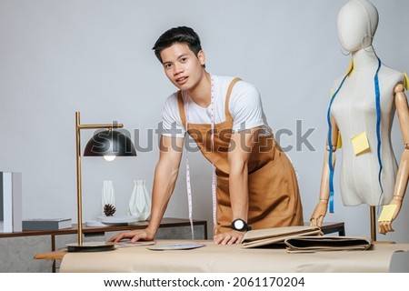 Portrait Young male fashion designer working with fabric and clothing sketches at the studio at home, full of tailoring tools, model and equipment on table