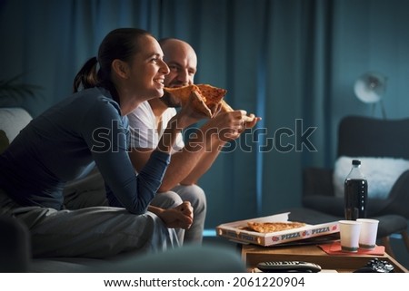 Happy couple relaxing at home together: they are sitting on the couch, watching TV and eating pizza Royalty-Free Stock Photo #2061220904