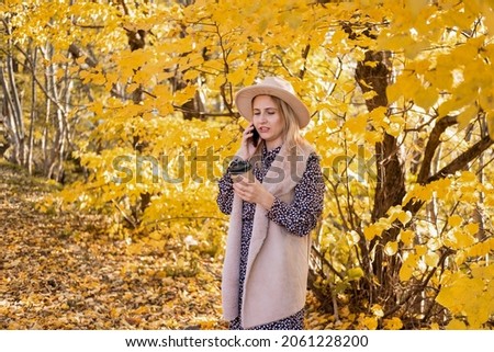 Young woman in fashionable clothes and hat drinking coffee, using phone in nature in autumn