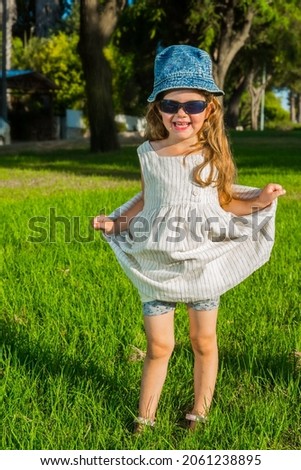 Adorable little girl with long blonde curls in a white sundress and panama. Warm summer day. Green lawn in the park. Girl smiles at the audience