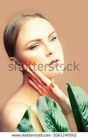 Glamour beauty shot of fashion model woman touching skin, palm leaf near face. Beige background. Skincare facial treatment health concept