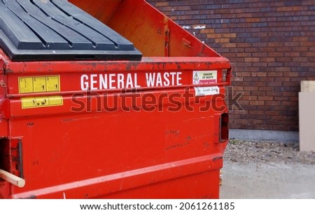 Closeup red industrial rubbish skip, left side of frame. Selective focus on front part of the metal bin with space to add text on blurry area, side of the bin. General waste, store clearance concept.