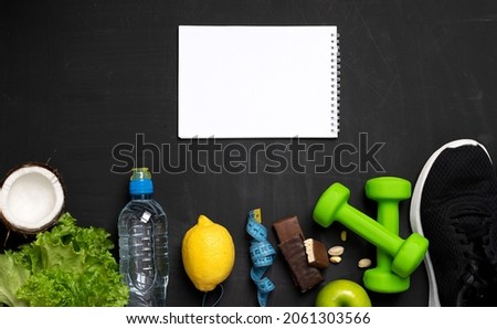 flat photo of sports equipment - dumbbells, table tennis rackets, tape measure, exercise equipment, water bottle and fruit, protein bars and a white tablet on a dark background for your recording.