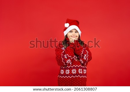 portrait of cute, Caucasian girl in red knitted Christmas sweater with reindeer holding palms to face, isolated on red background. 