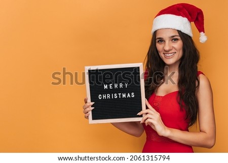 A smiling Caucasian woman in a red Santa hat holding a letter board with the inscription Merry Christmas on it. On a yellow isolated background. A place for your text.