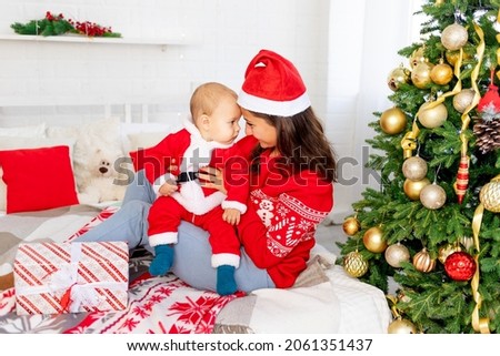 New Year or Christmas, a young mother with a baby on the bed at home by the Christmas tree in a Santa Claus costume hugging and waiting for the holiday smiling