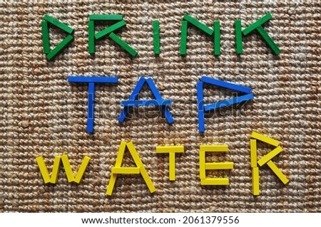 3D text in wooden bricks on jute carpet. It reads Drink Tap Water. Message for sustainability, climate change mitigation, climate action, carbon footprint reduction, lower CO2 emissions. Royalty-Free Stock Photo #2061379556