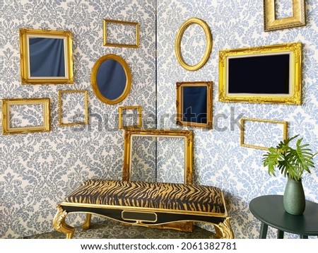 Multiple of golden vintage frames decorate on the wall with luxury wallpaper style and a tiger style cushion bench and a little round table with green leave in the vase.