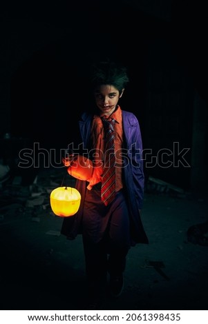 Serious boy with scary makeup in evil comic outfit looking at camera with spooky gaze while standing with glowing Jack O Lantern on black background in darkness during Halloween