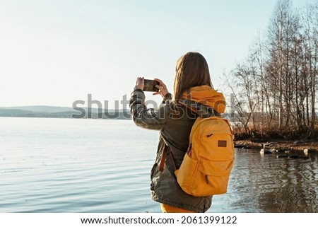 Rear view of young blonde woman with yellow backpack taking photo on smartphone of autumn landscape lake, using modern technology beauty in nature, fall season walking photographing