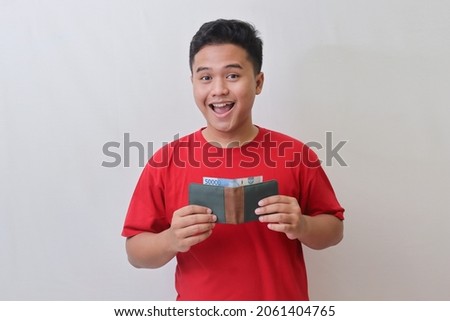 Portrait of attractive Asian man in red t-shirt looking at camera, holding a wallet with rupiah banknote. Isolated image on gray background Royalty-Free Stock Photo #2061404765