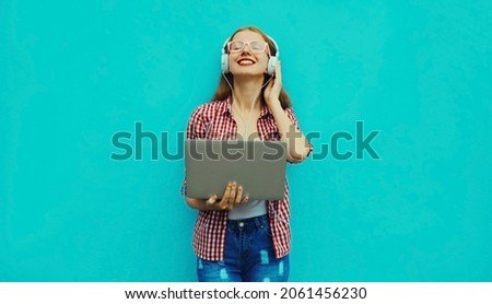 Portrait of modern young woman working with laptop listening to music in headphones on blue background