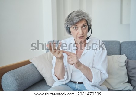 Serious senior woman teacher tutor wearing headset looking at web camera and explaining. Online conversation by videocall. Vlogger starting livestream, recording video for social media.