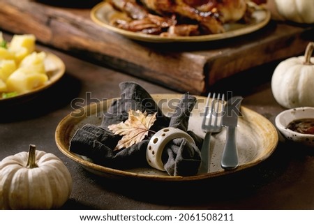 Holiday table setting with classic dishes roasted glazed duck with apples, boiled potatoes and sauce, empty ceramic plate with napkin and autumn leaf on dark table with autumn decor.