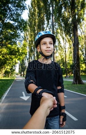 The boy is rollerblading on the track in the city park. The child learns to roller. An active walk with a child on the street. 
The child holds his mother's hand while rollerblading.
