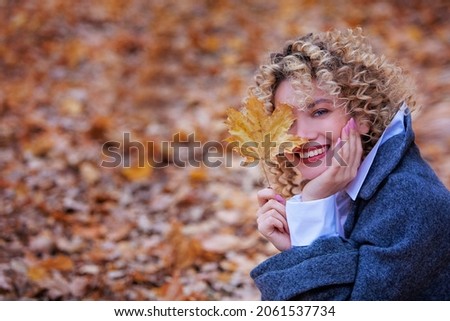 Young beautiful girl smiles and holds a yellow maple leaf in her hand. Girl has curly hair. Autumn Park. Copy space. Blurred background.