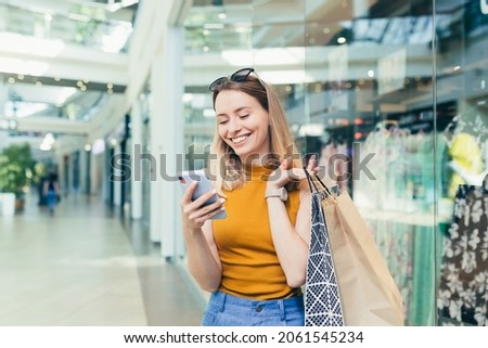Young woman consumer in the mall browses chat and uses using a smartphone. female standing with a mobile phone in her hands in shopping center. indoor. happy shopper girl with gift bags make purchases Royalty-Free Stock Photo #2061545234