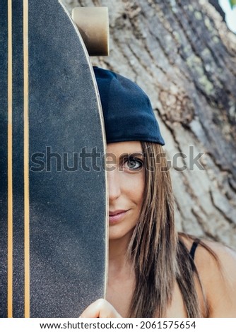 close-up detail of light-eyed Caucasian girl, hiding behind a skateboard. she has a cap and long hair. portrait of Caucasian girl with skateboard.