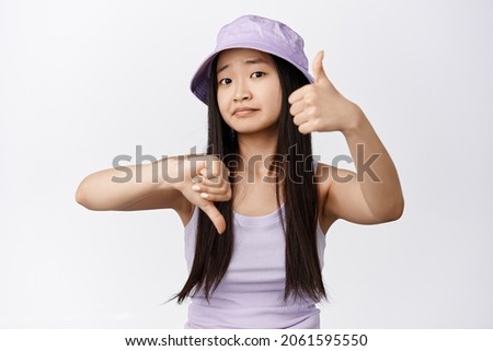 Cute asian girl asking your opinion, showing thumbs up and down and looking at camera, need advice, standing over white background