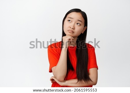 Thoughtful asian girl looking at upper left corner, thinking, making decision, standing in red t-shirt over white background