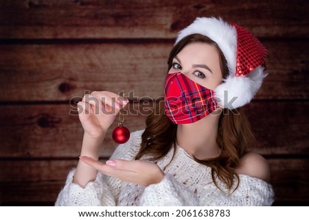 Woman wearing warm sweater and  protective mask holding Christmas red ball in hand on wooden background. COVID 19 constraint concept on Christmas.