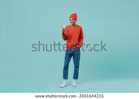 Full body young smiling happy african american man 20s in orange shirt hat use hold mobile cell phone chatting isolated on plain pastel light blue background studio portrait. People lifestyle concept Royalty-Free Stock Photo #2061644216